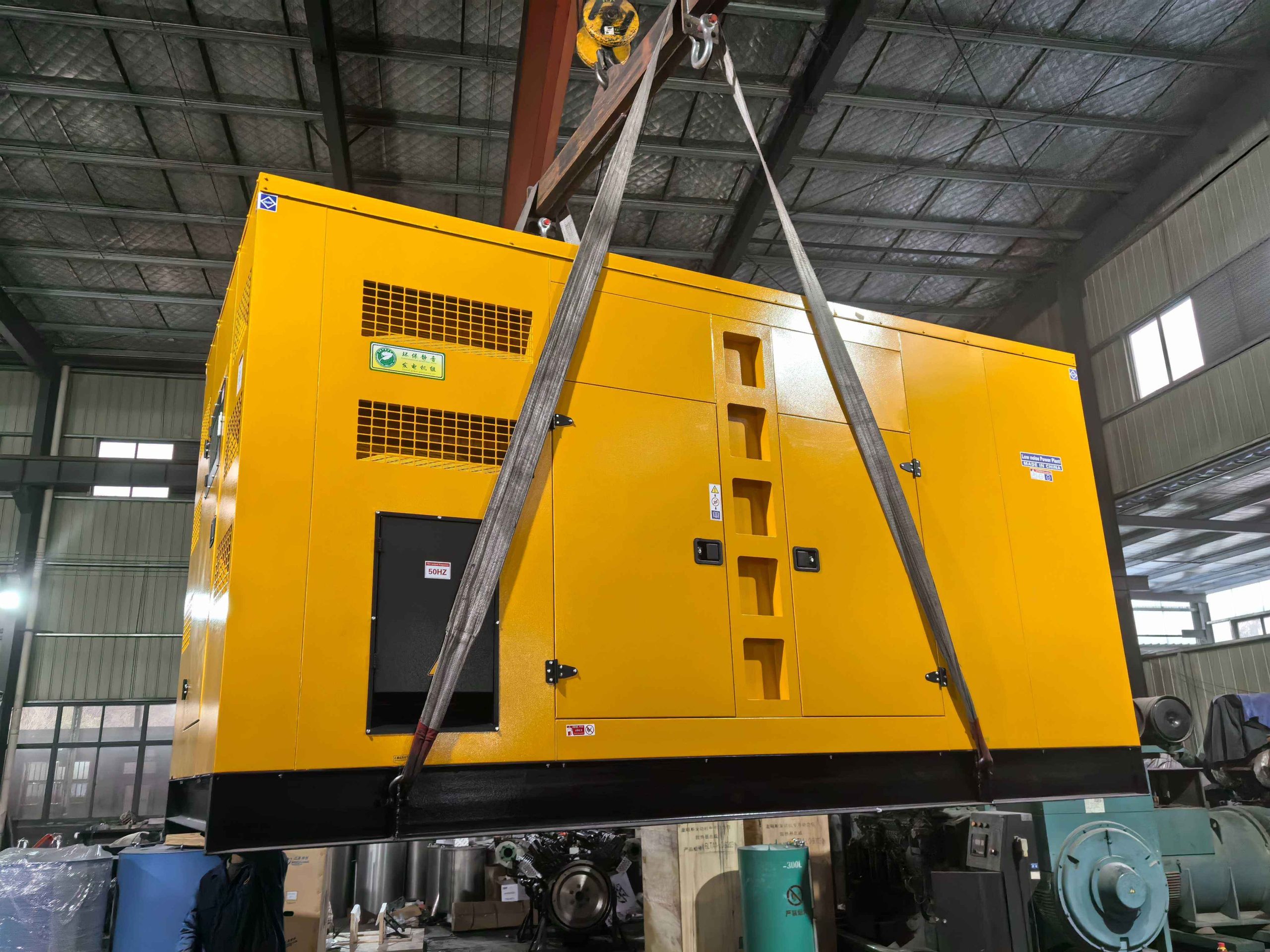 Yellow canopy diesel generator set lifted by crane slings in a workshop before dispatch