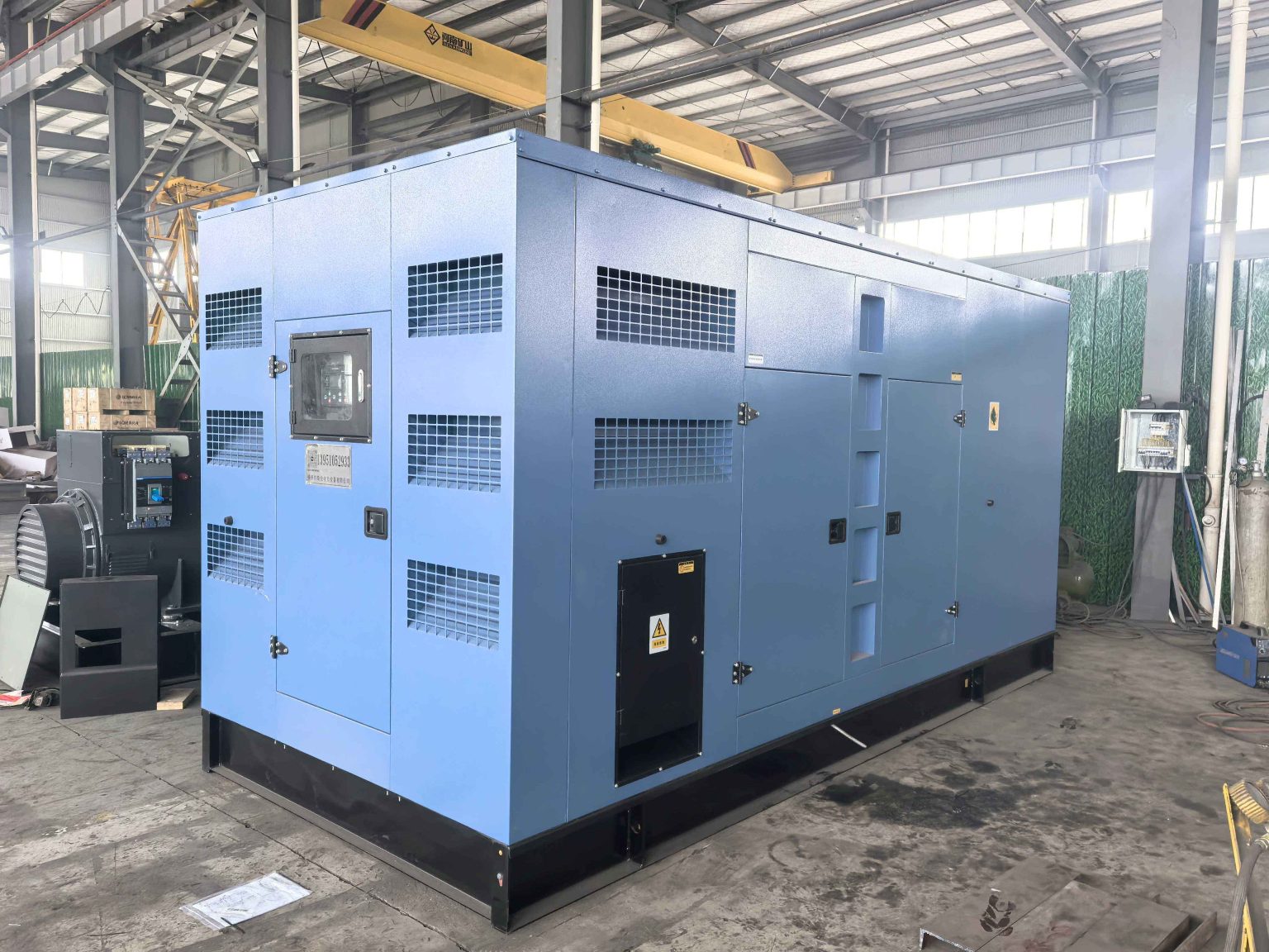Blue weatherproof generator canopy with a control panel window and ventilation louvres in an assembly workshop